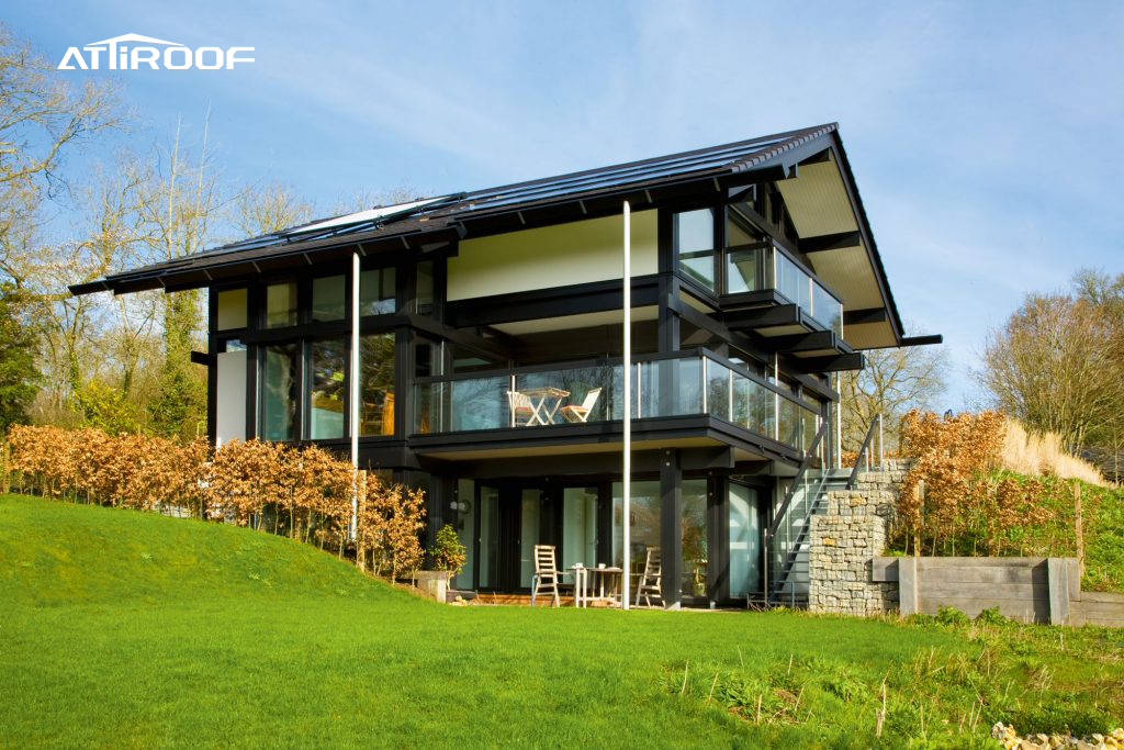 A newly built modern eco-friendly house with large glass windows and a sleek black synthetic resin tile roof, set against a natural landscape.